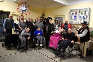 Duża grupa osób pozuje do wspólnego zdjęcia w pomieszczeniu udekorowanym świątecznie. Część uczestników siedzi na wózkach inwalidzkich, inni stoją obok; wszyscy znajdują się blisko siebie.