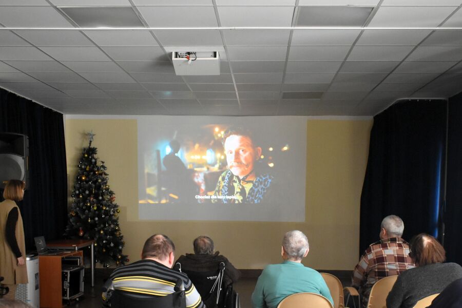 Kilka osób siedzi tyłem do aparatu i ogląda film wyświetlany na dużym ekranie w sali. Obok ekranu stoi udekorowana choinka, a pomieszczenie przypomina małą salę kinową.

