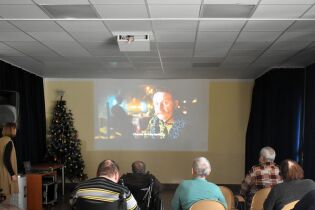 Kilka osób siedzi tyłem do aparatu i ogląda film wyświetlany na dużym ekranie w sali. Obok ekranu stoi udekorowana choinka, a pomieszczenie przypomina małą salę kinową.

