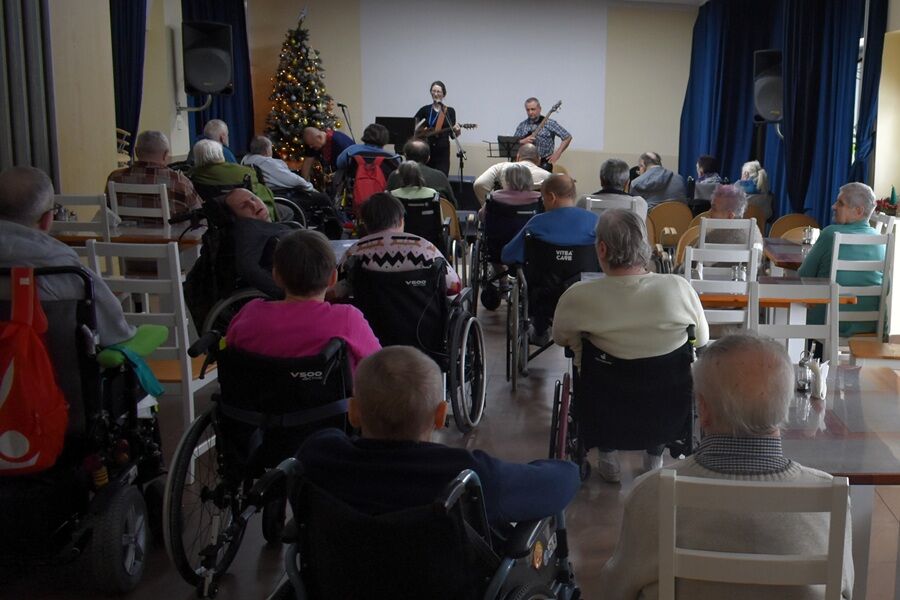 Grupa osób, w tym osoby poruszające się na wózkach inwalidzkich, siedzi na sali i słucha występu muzycznego z kolędami.
