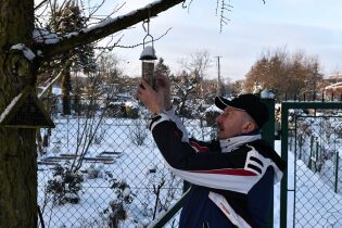 Mężczyzna w zimowej kurtce zawiesza karmnik dla ptaków na gałęzi drzewa w zaśnieżonym ogrodzie.

