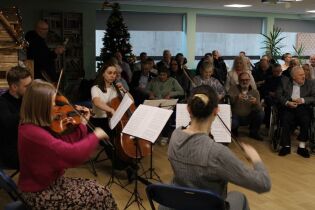 Koncert kwartetu smyczkowego w pomieszczeniu. Muzycy grają na instrumentach, a siedząca publiczność słucha występu. W tle choinka i świąteczne dekoracje.