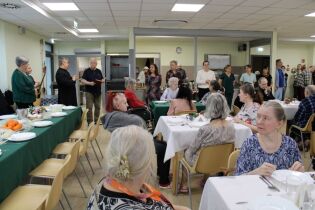 Grupa osób zgromadzona w sali. Część z nich siedzi przy stole, a inni stoją. W tle odbywa się przemówienie. Uczestnicy spotkania rozmawiają, a na stole znajdują się naczynia i jedzenie.