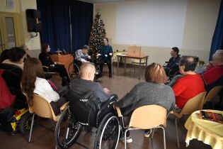 Grupa osób, w tym uczestnicy na wózkach inwalidzkich, siedzi w kręgu w sali. Prowadząca rozmawia z uczestnikami, a w tle stoi udekorowana choinka.