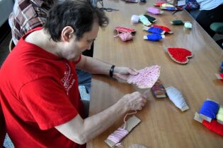 Osoba siedząca przy stole nawija kolorową włóczkę na kartonowe kształty. Na blacie leżą różne motki włóczki i gotowe prace plastyczne.