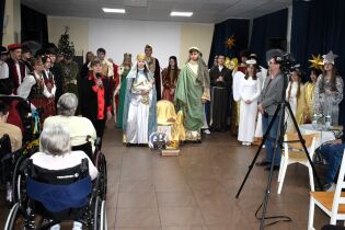 Grupa młodych ludzi w strojach jasełkowych stoi na scenie, przedstawiając scenę Bożego Narodzenia. Przed nimi siedzi publiczność, w tym osoby na wózkach, a z boku nagrywa kamerzysta.