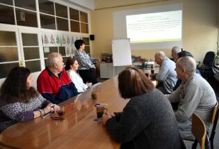 Grupa osób siedzi przy długim stole w sali spotkań i uważnie słucha prezentacji wyświetlanej na ekranie; prowadząca stoi obok flipcharta, a na stole widać kubki i notatki.