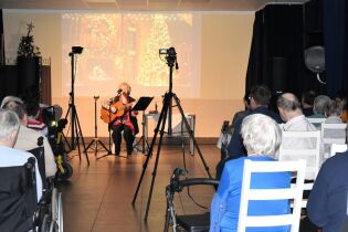 Kobieta siedząca na scenie gra na gitarze i śpiewa do mikrofonu podczas kameralnego koncertu; przed nią zgromadzona jest publiczność, część osób siedzi na wózkach inwalidzkich, scena jest oświetlona.