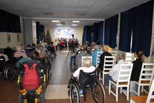 Publiczność zgromadzona w sali ogląda występ dzieci na scenie. Na ekranie za sceną widoczny jest napis z podziękowaniami oraz serca. Wiele osób siedzi na wózkach inwalidzkich, a atmosfera przypomina u