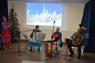 Czteroosobowy zespół muzyczny występuje w sali koncertowej. Muzycy grają na akordeonie, tubie, bębnie i skrzypcach. Obok stoi udekorowana choinka, a w tle wyświetlany jest zimowy krajobraz 
