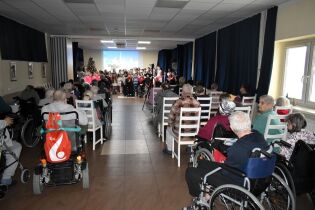 Widok sali z publicznością siedzącą na krzesłach i wózkach inwalidzkich, skierowaną w stronę sceny. Na scenie występuje grupa dzieci, a w tle widać choinkę i ekran z projekcją. 