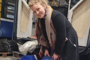 Kobieta w ciemnym płaszczu, z blond kręconymi włosami, pochyla się nad czarnym workiem z resztkami tkanin z tapicerek krakowskich tramwajów i autobusów, w hali MPK.