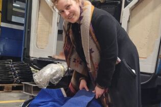 Kobieta w ciemnym płaszczu, z blond kręconymi włosami, pochyla się nad czarnym workiem z resztkami tkanin z tapicerek krakowskich tramwajów i autobusów, w hali MPK.