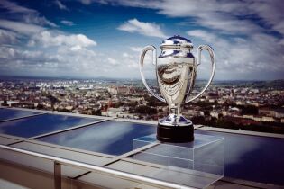 Srebrny duży Puchar IEM stoi na krawędzi oszklonego zadaszenia. W tle widoczna jest panorama Krakowa i pogodne niebieskie niebo z kilkoma białymi obłokami   