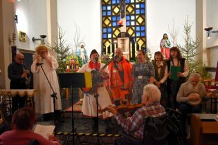 Wnętrze kościoła; grupa osób śpiewa kolędy przy ołtarzu, część gra na instrumentach, a publiczność siedzi na wózkach i ławkach.