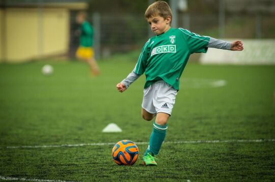 Grafika przedstawia małego chłopca w zielonej koszulce, przymierzającego się do strzelenia gola.