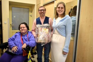 Trzy osoby stoją w pomieszczeniu; jedna z nich siedzi na wózku inwalidzkim. Uśmiechnięta grupa pozuje do zdjęcia, a osoba stojąca pośrodku trzyma zapakowany prezent.