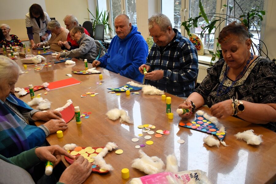 Grupa seniorów siedzi przy dużym stole i wspólnie wykonuje prace plastyczne, używając kolorowego papieru, kleju i waty. Zajęcia odbywają się w jasnej sali z oknami.