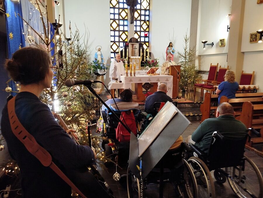 Wnętrze kościoła podczas nabożeństwa. Osoby na wózkach inwalidzkich uczestniczą we mszy, a w tle widać ołtarz, duchownych oraz świąteczne dekoracje z choinkami.