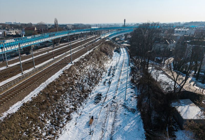 Widać prace remontowe nad ścieżką pieszo-rowerową przy Parku Kolejowy. Jest pochmurny dzień. Widać śnieg na inwestycji oraz tory kolejowe i okoliczne drzewa i zabudowania.