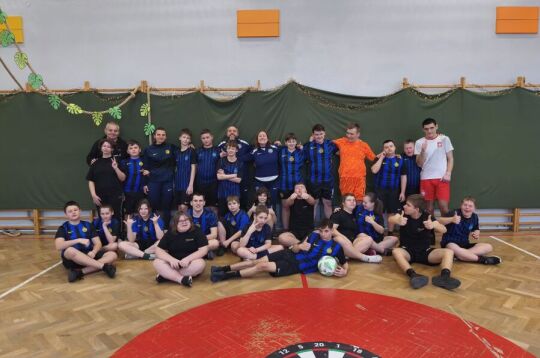 Projekt Inter Campus w Krakowie. Zdjęcie grupowe kilkunastu młodych osób oraz kilkorga dorosłych. Stoją w dwóch rzędach po drabinkami w sali gimnastycznej w koszulkach sportowych. 