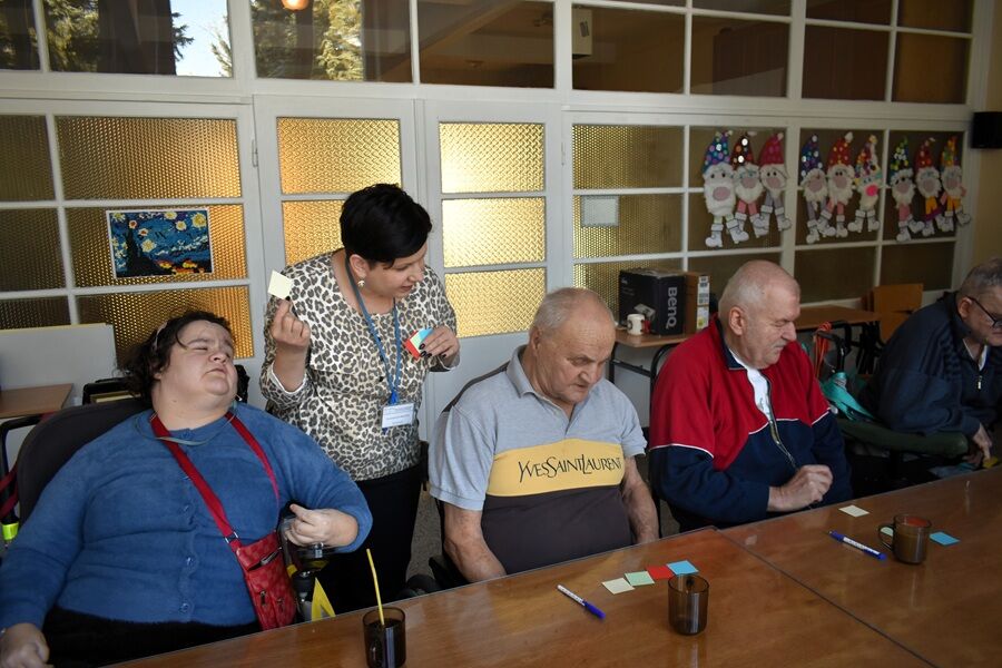 Seniorzy siedzą przy stole i wykonują proste zadania z użyciem kolorowych karteczek oraz długopisów. Prowadząca pochyla się nad jednym z uczestników, tłumacząc ćwiczenie. Atmosfera jest spokojna i sku