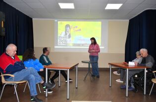 Kobieta stoi przed ekranem z wyświetloną prezentacją i przemawia do grupy seniorów siedzących przy stołach. Uczestnicy słuchają, część robi notatki. Sala jest jasna i przystosowana do spotkań edukacyj