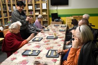 Grupa seniorów siedzi przy długim stole i bierze udział w zajęciach manualnych, formując małe figurki z jasnej masy plastycznej. Instruktorka stoi obok i pomaga uczestnikom. Na stole leżą tacki z mate