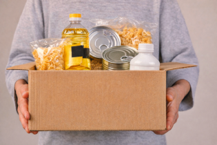 Osoba stojąca trzyma w rękach karton wypełniony produktami spozywczymi