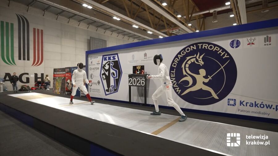 Dwóch zawodników walczy w pojedynku szermierki. Zawody odbywają się w hali AGH. Na drugim planie widać wiele osób oglądających rywalizację.