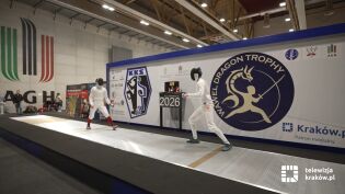 Dwóch zawodników walczy w pojedynku szermierki. Zawody odbywają się w hali AGH. Na drugim planie widać wiele osób oglądających rywalizację.