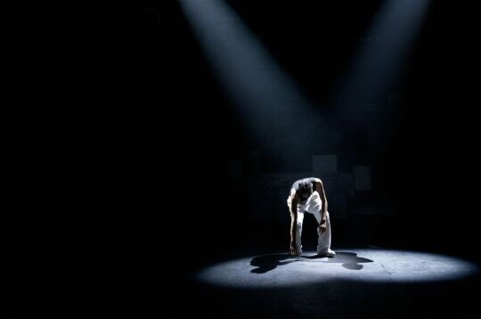 Grafika przedstawia pochyloną postać na scenie, oświetloną strumieniem światła z góry.