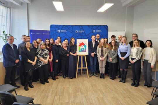 grupa osób stoi w kilku rzędach. po środku stoi na sztaludze logo centrum dla rodzin w formie kolorowego domku.