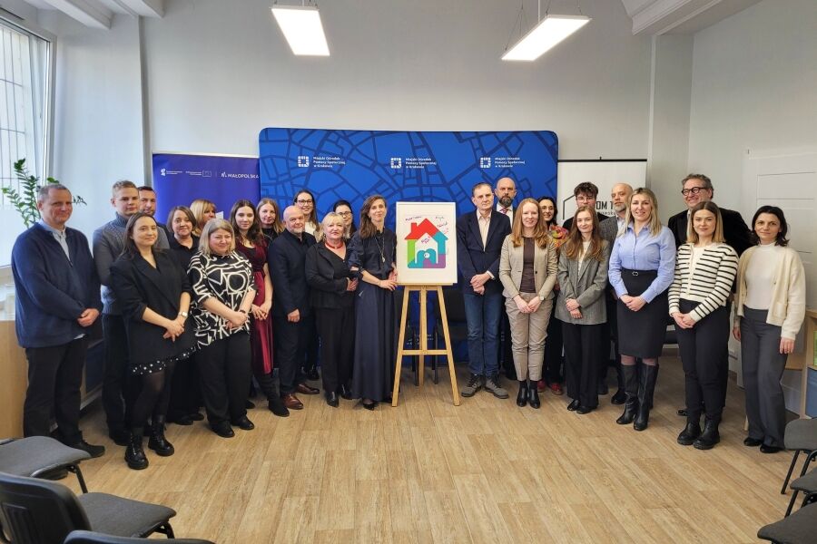 grupa osób stoi w kilku rzędach. po środku stoi na sztaludze logo centrum dla rodzin w formie kolorowego domku.