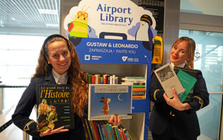 Dwie uśmiechnięte kobiety w uniformach lotniskowych stoją przed regałem z książkami napisem Odlotowa Biblioteka i trzymają w dłoniach książki w różnych formatach w językach obcych.      