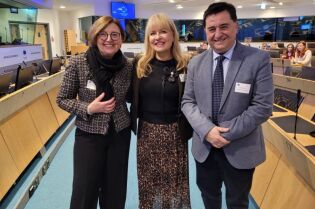 W sali konferencyjnej frontem do patrzącego stoją trzy osoby dwie kobiety i mężczyzna. Kobieta stojąca pośrodku trzyma oprawiony w ramkę certyfikat.   