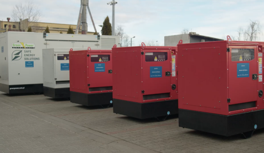 szereg metalowych dużych prostokątnych agregatów prądotwórczych stoi na betonowym podłożu w tle budynek przemysłowy. 