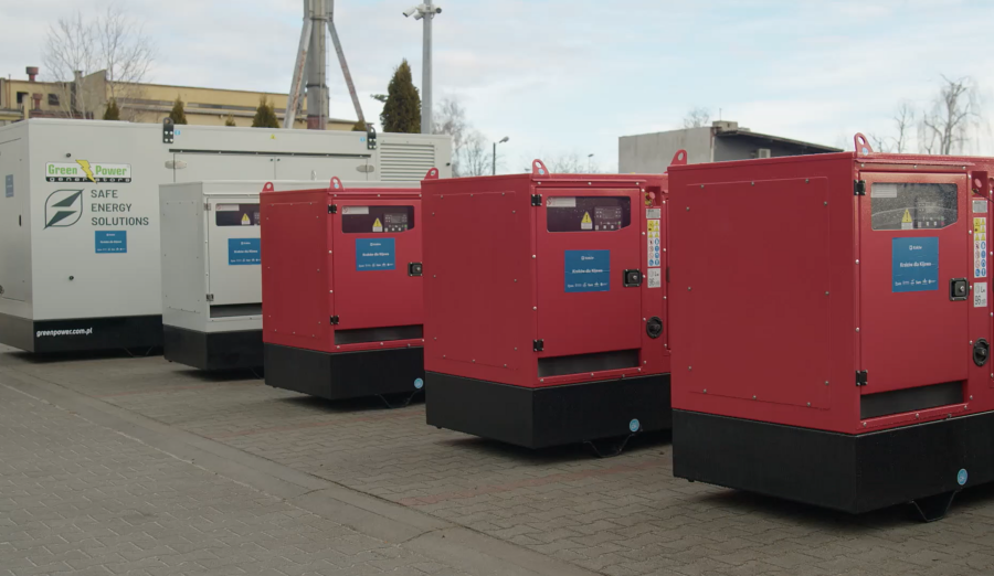 szereg metalowych dużych prostokątnych agregatów prądotwórczych stoi na betonowym podłożu w tle budynek przemysłowy. 