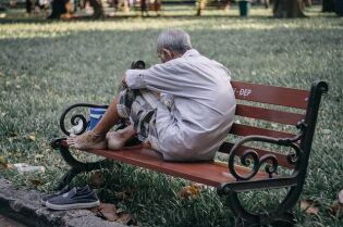 Grafika przedstawia postać, siedzącą na ławce, bez obuwia.