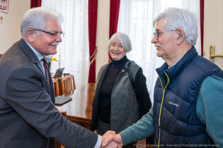 François Daubin po prawej stronie wita się przez podanie ręki z Zastępca Prezydenta Krakowa Stanisławem Kracikiem w jego gabinecie w tle stoi uśmiechnięta kobieta z siwymi włosami.         