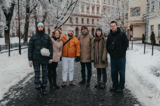 Fotografia przedstawia sześć osób trzy kobiety i trzech mężczyzn. Wszyscy są uczestnikami programu rezydencjalnego w Krakowie. Osoby te są w zimowych ubraniach i stoją tyłem do ulicy.
