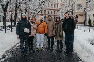 Fotografia przedstawia sześć osób trzy kobiety i trzech mężczyzn. Wszyscy są uczestnikami programu rezydencjalnego w Krakowie. Osoby te są w zimowych ubraniach i stoją tyłem do ulicy.