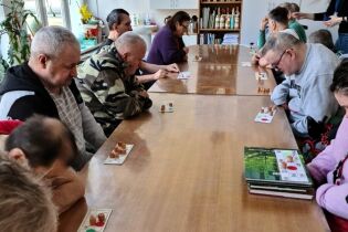 Grupa dorosłych osób siedzi wokół długiego stołu i uczestniczy w zajęciach stolikowych z użyciem plansz i drewnianych pionków. Uczestnicy są skupieni na zadaniach.