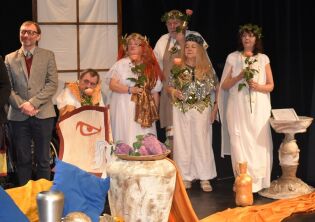 Kilka osób w kostiumach scenicznych stoi na scenie podczas przedstawienia teatralnego. Uczestnicy trzymają kwiaty i są ubrani w stylizowane stroje inspirowane antykiem.
