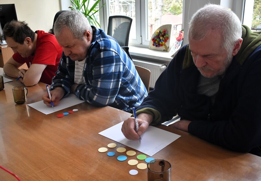 Trzech mężczyzn siedzi przy stole i wykonuje ćwiczenie polegające na zapisywaniu odpowiedzi na kartkach. Przed nimi leżą kolorowe żetony, a scena odbywa się w jasnym pomieszczeniu.