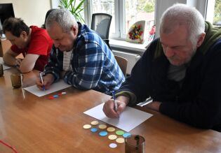 Trzech mężczyzn siedzi przy stole i wykonuje ćwiczenie polegające na zapisywaniu odpowiedzi na kartkach. Przed nimi leżą kolorowe żetony, a scena odbywa się w jasnym pomieszczeniu.