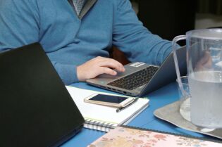 Grafika przedstawia fragment osoby, pracującej na laptopie, oraz telefon komórkowy, leżący na notatniku.