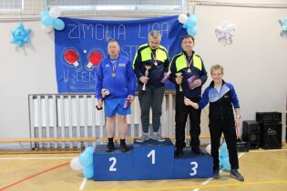 Trzech mężczyzn stoi na podium w sali gimnastycznej podczas dekoracji zwycięzców turnieju. Mają medale i trzymają puchary oraz nagrody. W tle na ścianie wisi baner „Zimowa Liga”