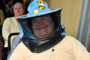 Uśmiechnięta kobieta siedzi w pomieszczeniu i ma na głowie kapelusz pszczelarski z siatką ochronną; w tle widoczna jest inna siedząca osoba oraz wnętrze budynku.