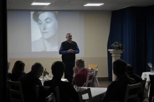 Mężczyzna stoi przed publicznością i przemawia, trzymając mikrofon; za nim na ekranie wyświetlony jest czarno-biały portret kobiety. Uczestnicy siedzą przy stołach w sali spotkań.